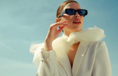 A woman in white jacket holding up her sunglasses.